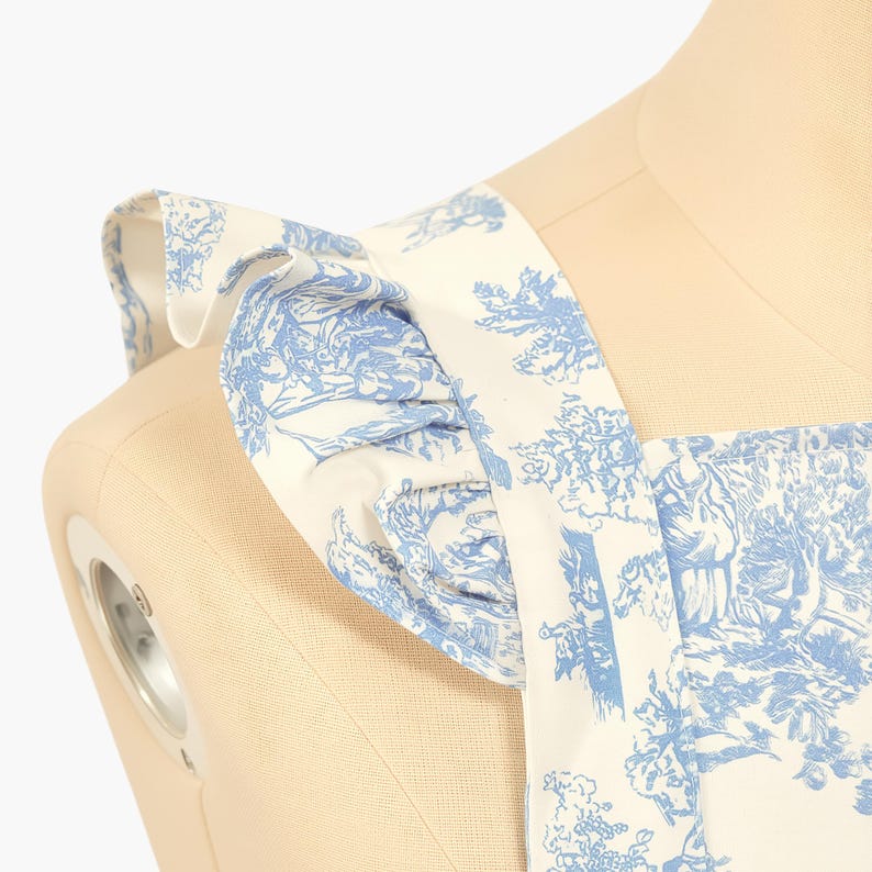 K&ouml;nnte beinhalten: Nahaufnahme eines wei&szlig;-blauen Toile-de-Jouy-Stoffes. Der Stoff zeigt eine detaillierte pastorale Szene mit B&auml;umen und Figuren. Das Kleidungsst&uuml;ck hat ger&uuml;schte Tr&auml;ger und wird auf einer Schaufensterpuppe pr&auml;sentiert.