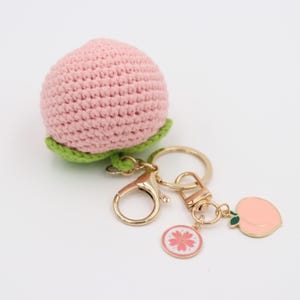 May include: A handmade peach keychain featuring a crocheted pink peach with green leaves. The keychain includes gold-colored hardware, a round charm with a pink flower design, and a peach-shaped charm.