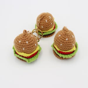 May include: Three handmade crochet burger keychains. The burgers feature brown buns with white seed details, yellow cheese, red tomato, and green lettuce. One keychain has a gold-colored clasp.
