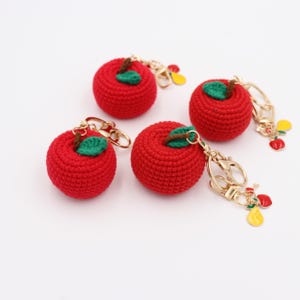 May include: Four red crocheted apple keychains with green leaves and gold-colored hardware. Each keychain has a small charm with red and yellow fruit. The apples are round and have a textured appearance.
