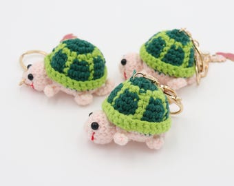 May include: Three handmade crochet turtle keychains. Each turtle has a pink body, a green and light green shell, and a gold-colored keychain ring. The turtles have black eyes and red mouths.