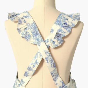 K&ouml;nnte beinhalten: Wei&szlig;er Sch&uuml;rze mit blauem Toile-de-Jouy-Muster, mit R&uuml;schen am Ausschnitt und gekreuzten Tr&auml;gern. Die Sch&uuml;rze wird auf einer Schaufensterpuppe gezeigt, die die R&uuml;ckansicht zeigt. Der Stoff hat ein detailliertes, klassisches Design.