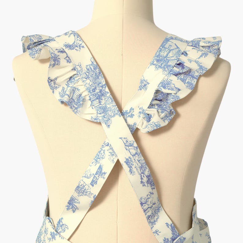 K&ouml;nnte beinhalten: Wei&szlig;er Sch&uuml;rze mit blauem Toile-de-Jouy-Muster, mit R&uuml;schen am Ausschnitt und gekreuzten Tr&auml;gern. Die Sch&uuml;rze wird auf einer Schaufensterpuppe gezeigt, die die R&uuml;ckansicht zeigt. Der Stoff hat ein detailliertes, klassisches Design.