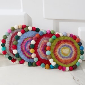 May include: A set of five colorful felt coasters. Each coaster features a circular design with concentric rings of various colors, and is trimmed with small, colorful pom-poms. The coasters are designed to protect surfaces from heat and moisture.
