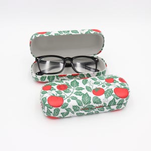Könnte beinhalten: Eine schwarze Brille liegt in einem offenen Etui mit Tomatenmuster. Ein zweites geschlossenes Etui mit dem gleichen Design befindet sich darunter. Die Etuis haben einen weißen Hintergrund mit roten Tomaten und grünen Blättern.