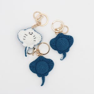 May include: Three handmade crochet stingray keyrings. One is white with a smiling face, and the other two are dark blue. Each keyring has a gold-coloured metal ring and clasp.