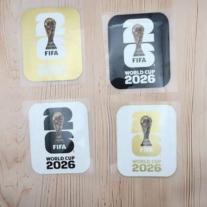May include: Four square stickers featuring the FIFA World Cup 2026 logo. Two stickers have a gold background with white text and graphics, while the other two have a black background with white text and graphics, all displaying the trophy icon.