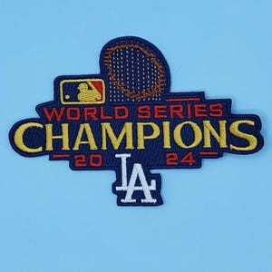 May include: Embroidered patch featuring the text "WORLD SERIES CHAMPIONS" in yellow, with a navy blue background and red accents. The patch includes the MLB logo and the text "20 24" with the LA logo. The patch is on a light blue background.
