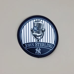 May include: A circular embroidered patch featuring a vintage-style microphone centered over a white and navy blue pinstripe background. The text JOHN STERLING is printed in white below the microphone, with the New York Yankees logo above and below.