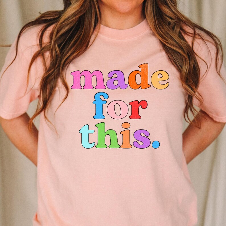 May include: A light pink t-shirt with the phrase "made for this" in colorful, bubble letters. The text is in shades of pink, orange, blue, red, green, and purple. The shirt has a crew neck and short sleeves.