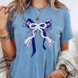 May include: A light blue t-shirt featuring a large bow design. The bow has a gingham pattern in shades of blue and is adorned with a string of white pearls. The shirt is a casual style, perfect for everyday wear.