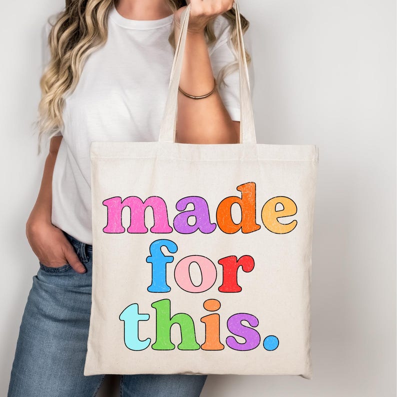 May include: Cream-colored tote bag with the words "made for this" in colorful, bubble letters. The bag is held by a person wearing a white t-shirt and blue jeans. The text is in pink, orange, purple, blue, red, green, and blue.
