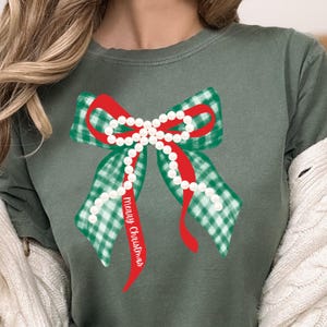 Puede incluir: Camiseta verde salvia con un diseño de lazo navideño. El lazo tiene un patrón de cuadros verdes y blancos, una cinta roja y detalles de perlas blancas. El texto "Merry Christmas" está impreso en la cinta.