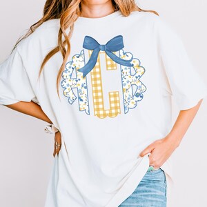 May include: White t-shirt featuring a floral and gingham monogram design with a blue bow. The monogram includes the letter "C" in a yellow gingham pattern, surrounded by a scalloped edge with blue and yellow floral accents. The bow is a solid blue color.