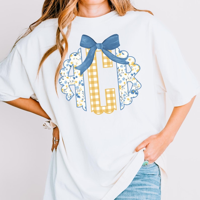 May include: White t-shirt featuring a floral and gingham monogram design with a blue bow. The monogram includes the letter "C" in a yellow gingham pattern, surrounded by a scalloped edge with blue and yellow floral accents. The bow is a solid blue color.