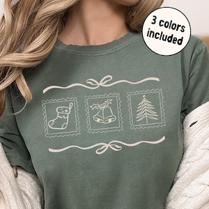 May include: Sage green t-shirt with three Christmas-themed postage stamp designs: a stocking, bells, and a Christmas tree. The text "3 colours included" is in a circle in the upper right.