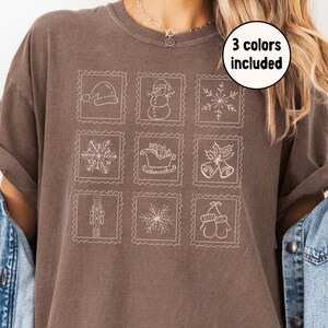 May include: A brown t-shirt featuring nine white Christmas-themed postage stamp designs. The stamps include a Santa hat, snowman, snowflakes, sleigh, bells, nutcracker, and mittens. The text "3 colors included" is in a white circle.