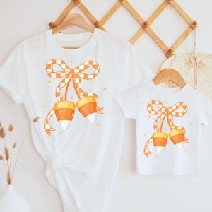 Może przedstawiać: Białe koszulki z wzorem cukierków kukurydzianych. Wzór przedstawia pomarańczową kokardę w kratkę z akcentami cukierków kukurydzianych. Koszulki są prezentowane na drewnianych wieszakach na neutralnym tle.