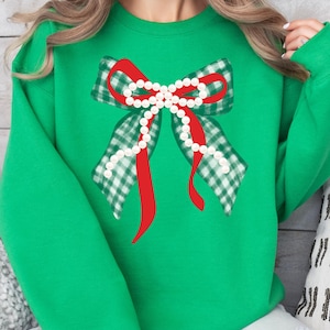 Puede incluir: Una sudadera verde con un diseño de lazo grande. El lazo tiene un centro rojo y alas a cuadros verdes y blancos, adornadas con una hilera de perlas blancas. La sudadera es de color verde liso.