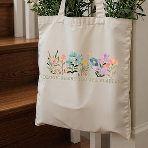 K&ouml;nnte beinhalten: Cremefarbene Tragetasche mit floralem Design und dem Text "BLOOM WHERE YOU ARE PLANTED" in Gr&uuml;n. Das florale Design weist verschiedene Farben auf, darunter Blau, Orange und Lila. Die Tasche ist mit gr&uuml;nen Pflanzen gef&uuml;llt.