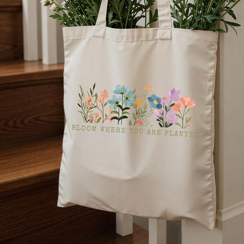 K&ouml;nnte beinhalten: Cremefarbene Tragetasche mit floralem Design und dem Text "BLOOM WHERE YOU ARE PLANTED" in Gr&uuml;n. Das florale Design weist verschiedene Farben auf, darunter Blau, Orange und Lila. Die Tasche ist mit gr&uuml;nen Pflanzen gef&uuml;llt.