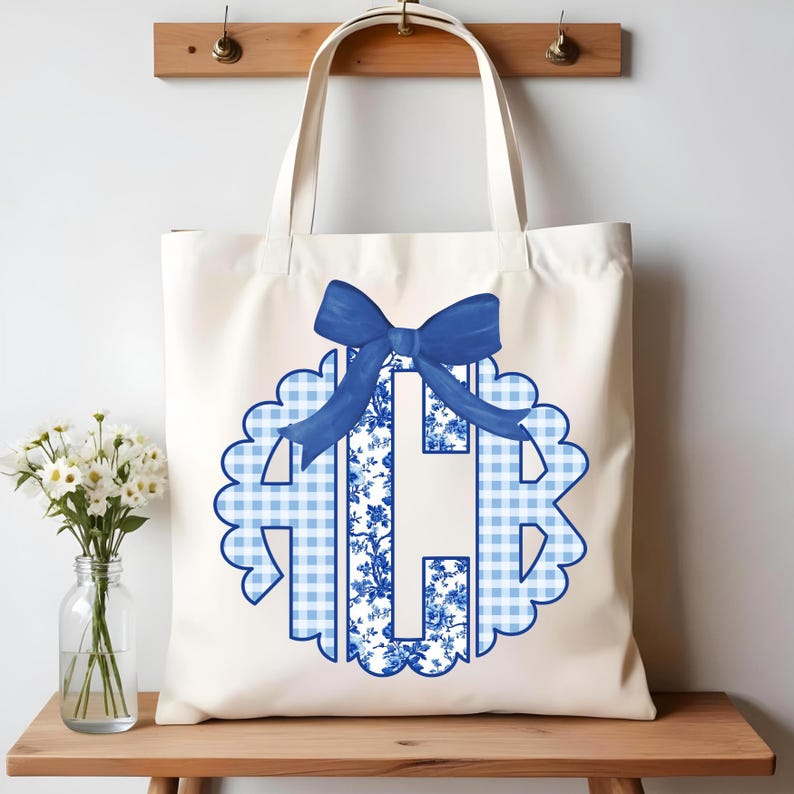 May include: Cream tote bag with a blue monogram featuring a scalloped edge. The monogram includes a blue bow and a combination of floral and gingham patterns. The bag has two handles and is displayed on a wooden surface.