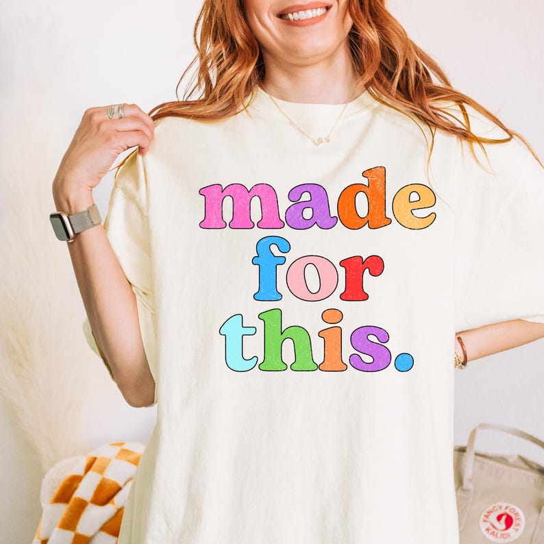 May include: Cream-colored t-shirt with the phrase "made for this" in colorful bubble letters. The text is in pink, purple, orange, blue, red, and green. The shirt is a casual, everyday wear item.