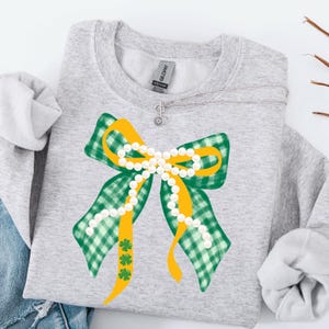 May include: A light grey sweatshirt with a large bow design. The bow features green and white checked ribbons, yellow accents, and a string of white pearls. Three green shamrocks are below the bow. A silver necklace with a flower pendant is also visible.