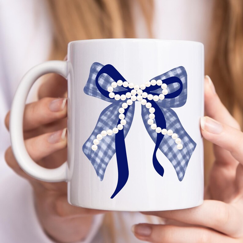 May include: White ceramic mug featuring a blue bow design. The bow has a gingham pattern and is accented with a dark blue ribbon and a string of white pearls. The mug has a white handle.