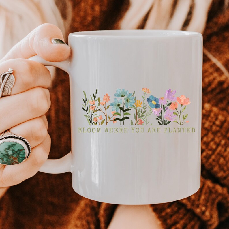 K&ouml;nnte beinhalten: Wei&szlig;er Keramikbecher mit einem bunten Blumendesign. Er zeigt blaue, rosa und lila Blumen. Der Becher tr&auml;gt den gr&uuml;nen Text "BLOOM WHERE YOU ARE PLANTED". Ein sch&ouml;nes Geschenk f&uuml;r Blumenliebhaber.