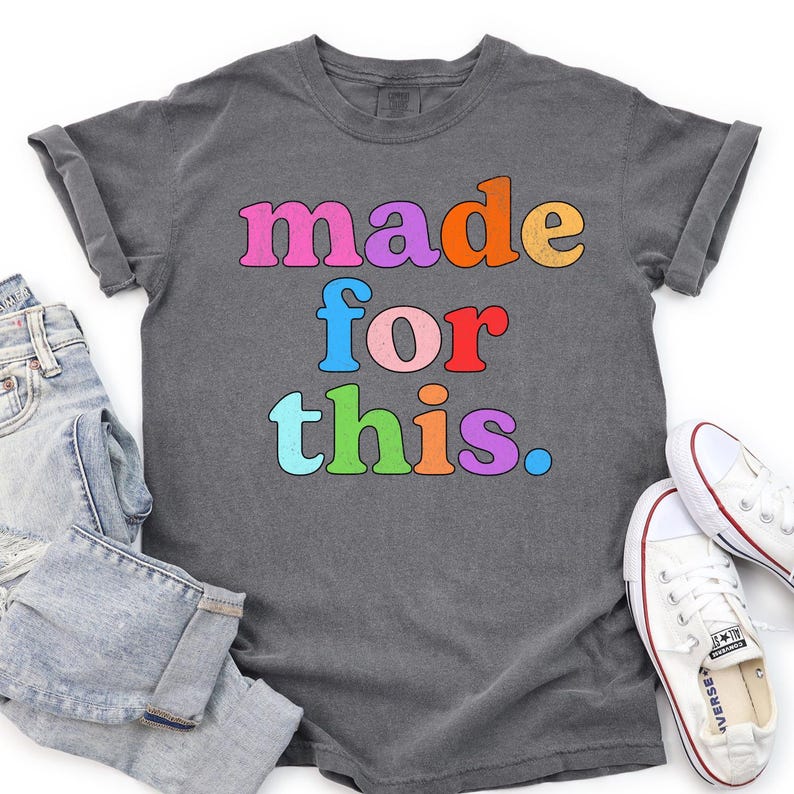 May include: Dark gray t-shirt with the words "made for this" in colorful, retro-style lettering. The shirt is paired with blue jeans and white sneakers, creating a casual, everyday look. The shirt's design is eye-catching and bold.
