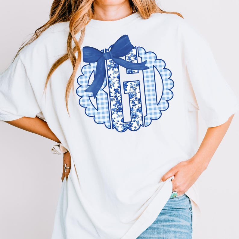 May include: White t-shirt with a blue bow and monogram design. The monogram features a blue floral pattern within a scalloped border, set against a blue and white gingham background. The shirt has a loose fit.