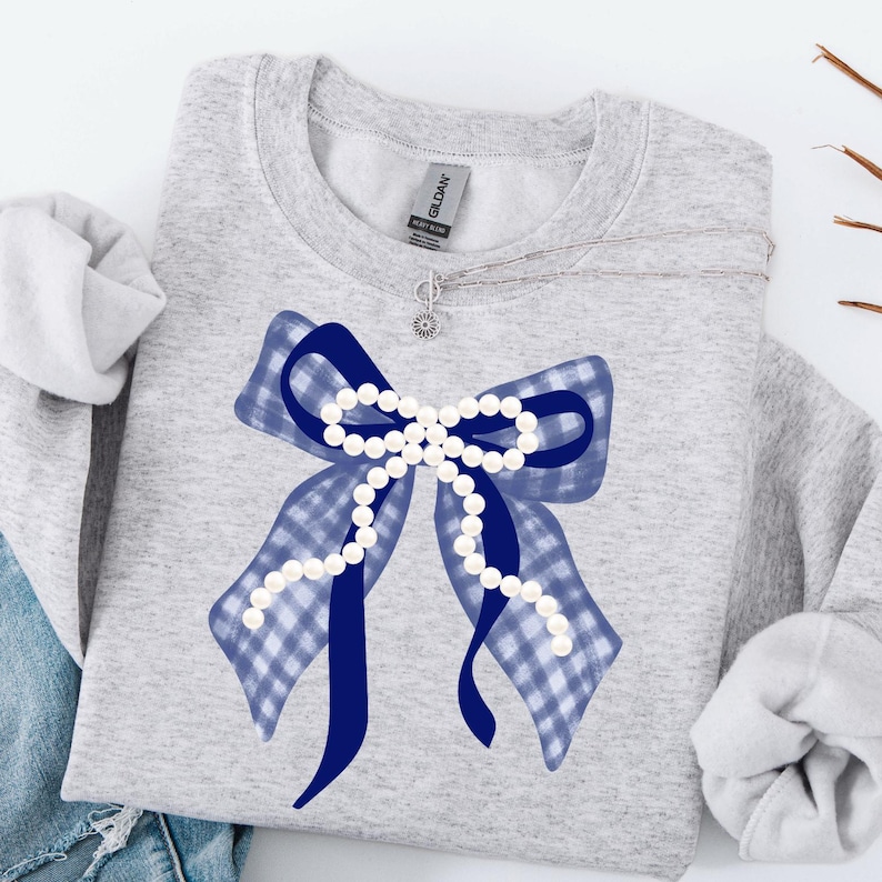 May include: A light gray sweatshirt featuring a large bow design. The bow is a combination of a blue gingham pattern and solid blue ribbons, adorned with a string of white pearls. A silver necklace with a small charm is visible.