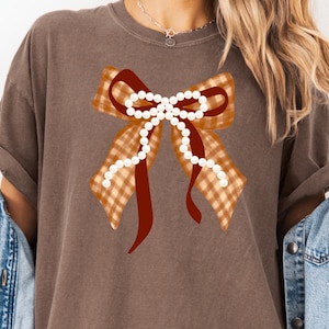 Op de afbeelding: Bruin t-shirt met een groot strikdesign. De strik heeft een ruitpatroon in bruin- en oranje tinten, een donkerrood midden en parelaccenten. Het shirt heeft een effen bruine kleur.