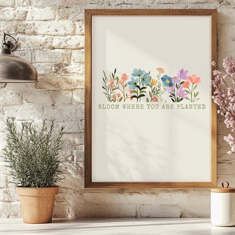 K&ouml;nnte beinhalten: Ein gerahmter Kunstdruck mit einer Aquarellillustration von bunten Blumen in Rosa-, Blau- und Lilat&ouml;nen. Der Text "BLOOM WHERE YOU ARE PLANTED" ist darunter in Gr&uuml;n gedruckt. Der Rahmen ist aus Holz.