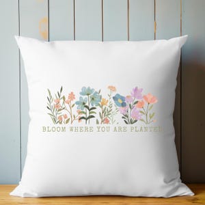 K&ouml;nnte beinhalten: Wei&szlig;es quadratisches Kissen mit einem Aquarell-Blumenmuster in Rosa-, Blau- und Lilat&ouml;nen. Das Kissen tr&auml;gt den Text "BLOOM WHERE YOU ARE PLANTED" in hellgr&uuml;ner Schrift. Das Kissen liegt auf einer hellblauen Holzoberfl&auml;che.