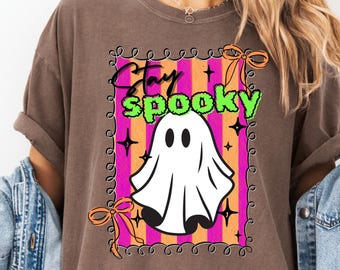May include: Brown t-shirt with a Halloween-themed graphic. The graphic features a white ghost with the words "Stay Spooky" in black and green, set against a pink and orange striped background, framed with a decorative border and orange bows.