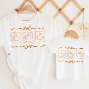 May include: White t-shirts with a pumpkin design. The adult shirt has a larger design than the child's shirt. Each shirt features three pumpkin stamps in a row, with a decorative line above and below.