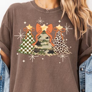 Puede incluir: Camiseta marrón con tres árboles de Navidad con diseños únicos: un árbol a cuadros verdes y blancos, un árbol de camuflaje y un árbol con estampado de leopardo. Cada árbol tiene una estrella en la parte superior y está adornado con un lazo naranja. Estrellas blancas rodean los árboles.