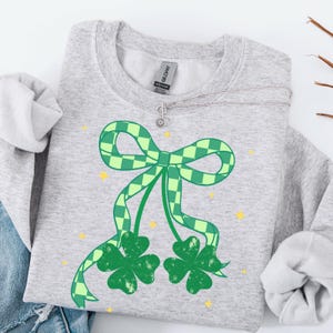 May include: A light gray sweatshirt featuring a large green bow with a checkered pattern and two green shamrocks. The design includes yellow star accents. The sweatshirt has a crew neck and long sleeves.