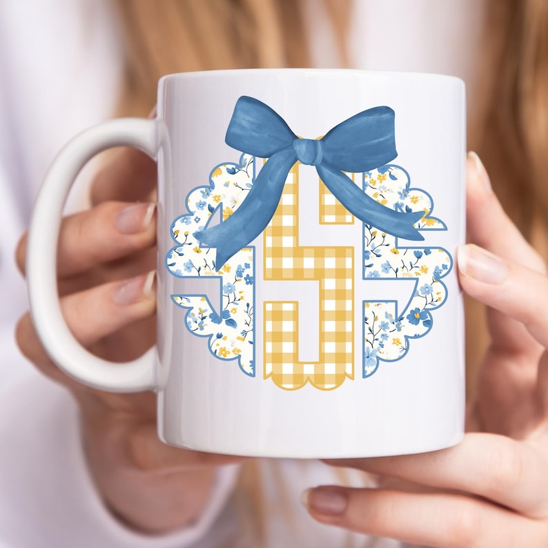 May include: White ceramic mug featuring a blue bow and a floral and gingham design. The design includes a blue bow at the top, with a floral pattern on the left and a yellow gingham pattern on the right. The mug is held by a person.