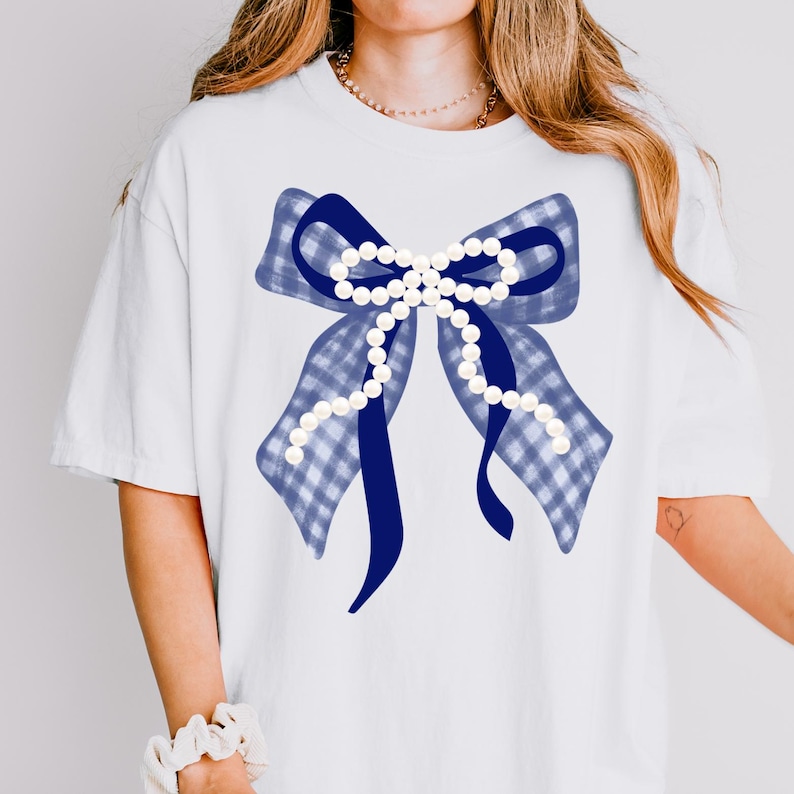 May include: White t-shirt featuring a large blue bow with a gingham pattern and a solid blue center. A string of white pearls adorns the center of the bow. The shirt is a casual style.