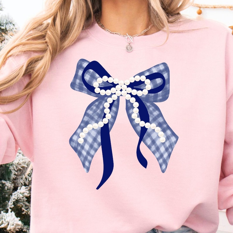 May include: Pink sweatshirt featuring a large blue bow with a gingham pattern and a dark blue ribbon. The bow is adorned with a string of white pearls. The sweatshirt is a solid light pink color.