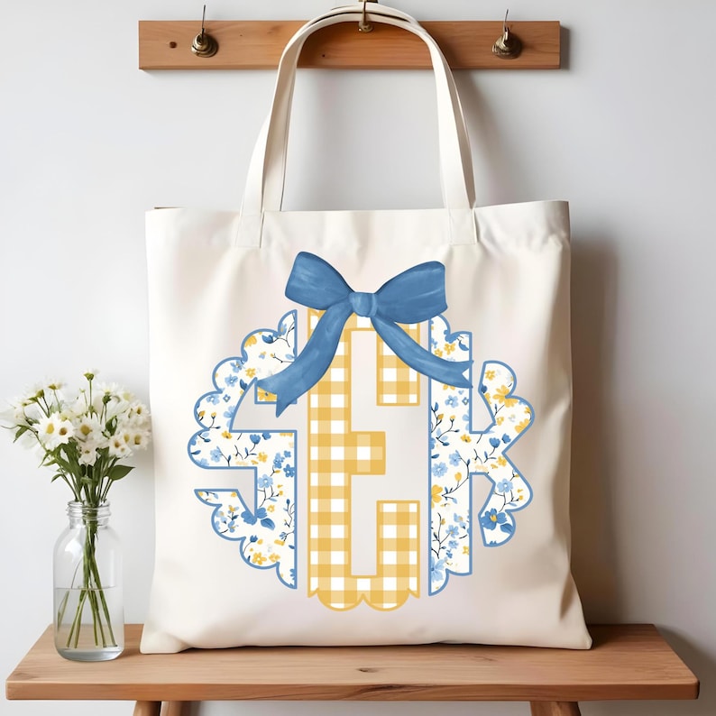 May include: Cream-colored tote bag featuring a monogram design with a blue bow. The monogram incorporates yellow gingham and floral patterns with blue flowers. The bag has a simple design with long handles for carrying.