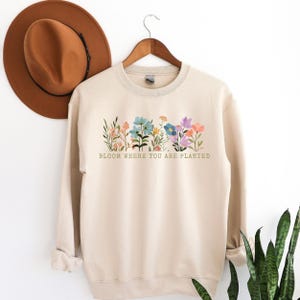 K&ouml;nnte beinhalten: Beiges Sweatshirt mit einem Blumendesign mit Blumen in verschiedenen Farben und gr&uuml;nen Bl&auml;ttern. Der Text "BLOOM WHERE YOU ARE PLANTED" ist unter den Blumen aufgedruckt. Ein brauner Hut h&auml;ngt im Hintergrund.