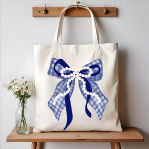 May include: Cream-colored tote bag featuring a large blue bow with a gingham pattern and a string of white pearls. The bow is centered on the bag, with long, flowing ribbons. The bag has two handles and is displayed on a wooden surface.