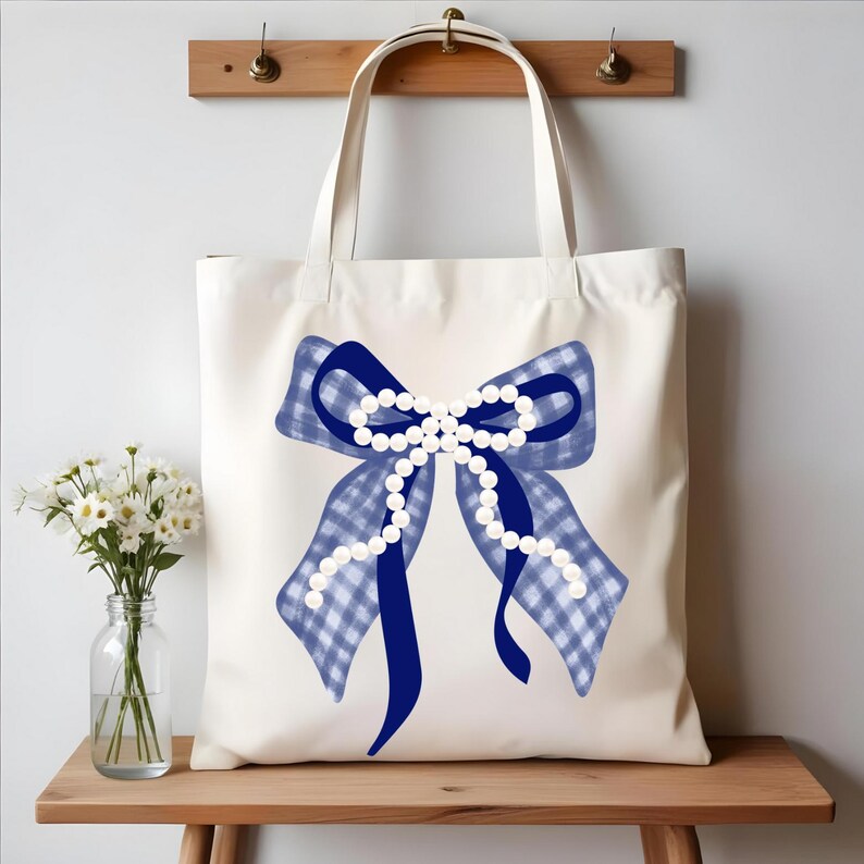 May include: Cream-colored tote bag featuring a large blue bow with a gingham pattern and a string of white pearls. The bow is centered on the bag, with long, flowing ribbons. The bag has two handles and is displayed on a wooden surface.