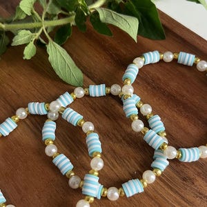 May include: Three bracelets featuring a repeating pattern of light blue and white striped disc beads, interspersed with small metallic gold spheres and round white faux pearls, arranged in a circular formation on a wooden surface.