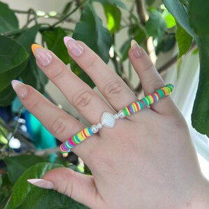 Puede incluir: Una pulsera colorida con una serie de cuentas redondas y planas de arcoíris. La pulsera está adornada con un dije de concha blanca y pequeñas cuentas blancas redondas. La pulsera se lleva en una mano con uñas cuidadas.