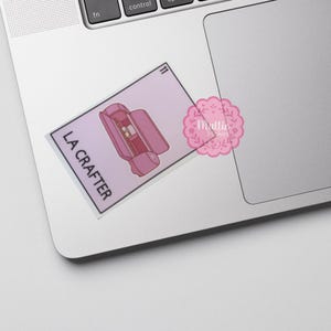 以下が含まれることがあります： クラフトツールケースのピンク色のイラストと黒色の「LA CRAFTER」の文字が描かれたステッカー。数字の「11」が右上隅にあります。ピンク色の花のロゴが右側にあります。